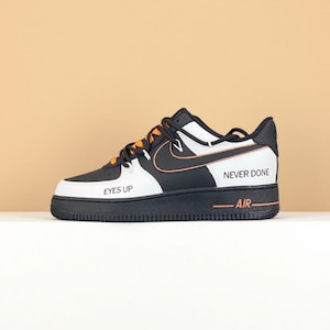 May include: Black, white, and orange Nike Air Force 1 trainer. The shoe features the words "EYES UP" on the toe, "NEVER DONE" on the side, and "AIR" on the midsole. The Nike swoosh is orange.