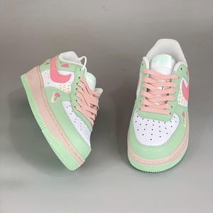 May include: Pair of pastel-colored sneakers. The shoes feature a white base with mint green and pink accents. The design includes pink laces, a pink swoosh, and a mint green sole. Small mushroom graphics are visible on the side.