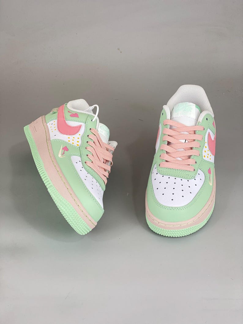 May include: Pair of pastel-colored sneakers. The shoes feature a white base with mint green and pink accents. The design includes pink laces, a pink swoosh, and a mint green sole. Small mushroom graphics are visible on the side.