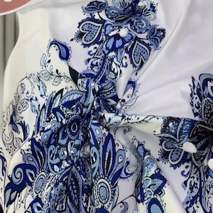 K&ouml;nnte beinhalten: Wei&szlig;er Stoff mit blauem Blumenmuster. Das Design zeigt komplizierte Paisley- und Blumenmotive in Blaut&ouml;nen, die einen detaillierten und eleganten Look erzeugen. Der Stoff ist gerafft und geknotet, was die Kontinuit&auml;t des Musters zeigt.