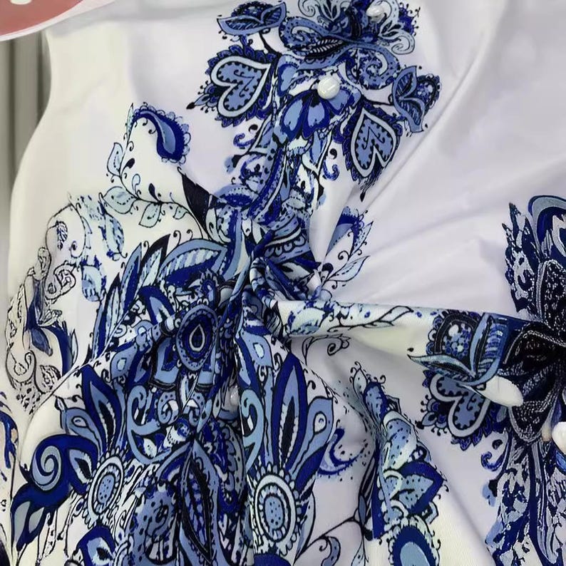 K&ouml;nnte beinhalten: Wei&szlig;er Stoff mit blauem Blumenmuster. Das Design zeigt komplizierte Paisley- und Blumenmotive in Blaut&ouml;nen, die einen detaillierten und eleganten Look erzeugen. Der Stoff ist gerafft und geknotet, was die Kontinuit&auml;t des Musters zeigt.