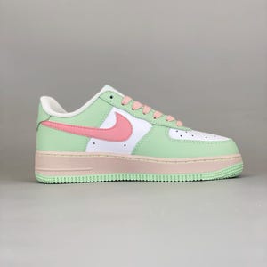 May include: A pastel-colored Nike Air Force 1 sneaker. The shoe features a white toe box, a light green mid-panel, and a pink swoosh. The sole is a light pink, and the laces are pink. The heel is white.
