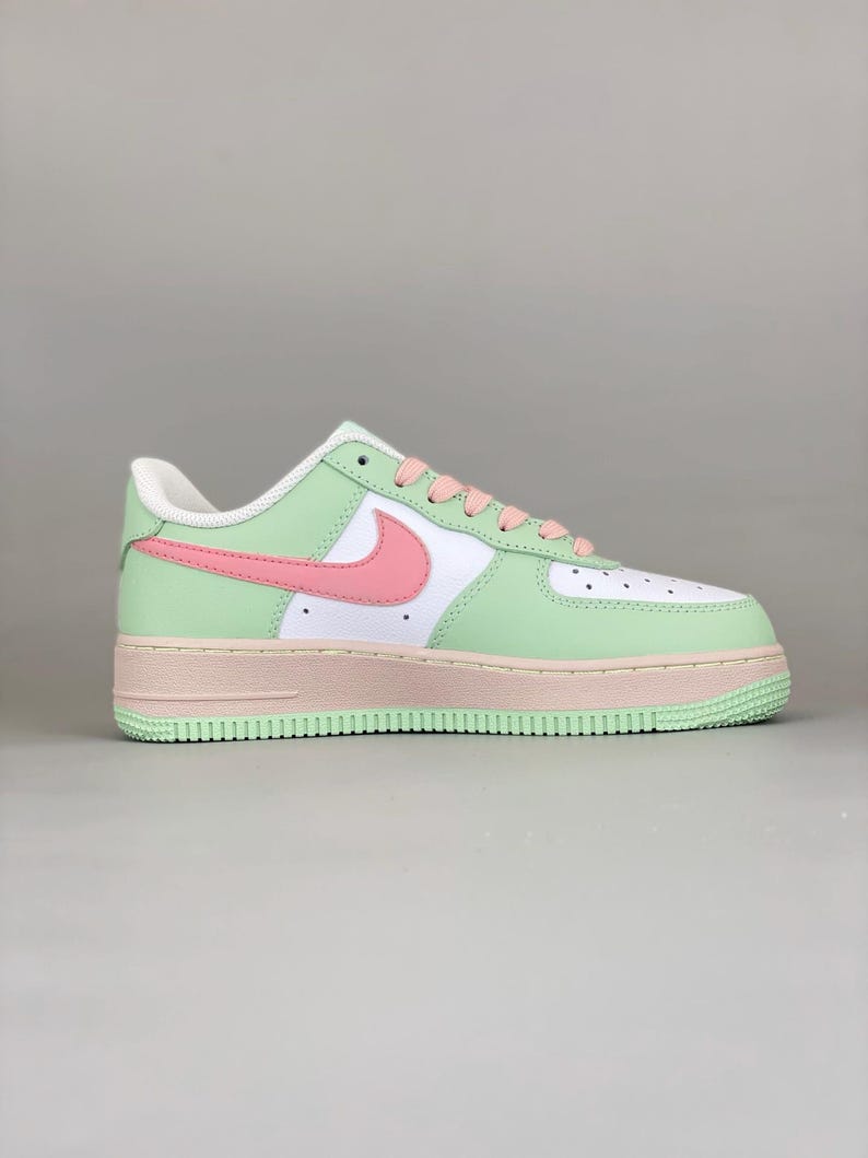 May include: A pastel-colored Nike Air Force 1 sneaker. The shoe features a white toe box, a light green mid-panel, and a pink swoosh. The sole is a light pink, and the laces are pink. The heel is white.