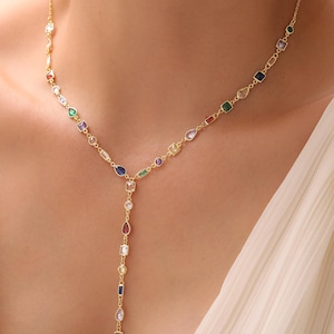 May include: A gold necklace with a Y-shaped pendant. The necklace has a fine chain decorated with colourful gemstones in various shapes, including round, oval, and rectangular. The gemstones are in shades of red, blue, green, purple, and clear.