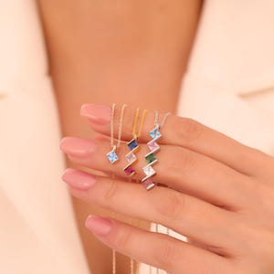 May include: A collection of delicate necklaces with small, colorful gemstone pendants. The pendants are square or rectangular, with a zig-zag design. The necklaces are displayed against a soft, neutral background, highlighting the vibrant colors of the gemstones.