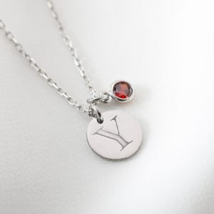 May include: A silver necklace with a small round pendant engraved with the letter "Y" and a small red gemstone. The necklace is on a white, textured background.