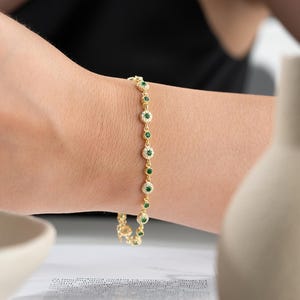 May include: A gold chain bracelet featuring a series of circular links, each set with a central round green gemstone surrounded by a halo of small clear white stones. The bracelet has a delicate metal construction and a secure clasp mechanism.