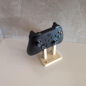 Può includere: Un controller wireless per videogiochi nero appoggiato su un supporto in plastica beige. Il supporto presenta una base rettangolare con la scritta XBOX e il logo circolare Xbox in rilievo sulla parte anteriore. Il controller ha due levette.