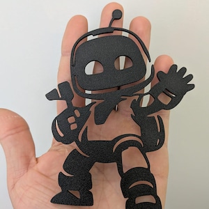 Op de afbeelding: Zwarte robotvormige uitsparing met een getextureerd oppervlak. De robot heeft een lachend gezicht, een helm en houdt een gitaar vast. De robot zwaait met één hand en heeft een beenontwerp met drie gebogen lijnen. De uitsparing wordt in een hand gehouden.