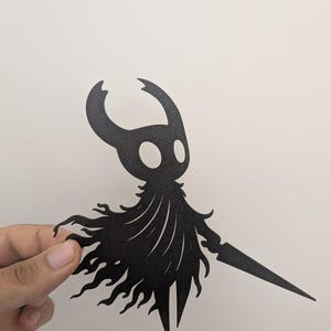 Puede incluir: Silueta negra de un personaje con cabeza con cuernos, ojos grandes y una capa fluida, sosteniendo una espada. El personaje está cortado de un material plano, probablemente metal o papel grueso, y es sostenido por una mano sobre un fondo blanco.