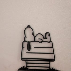 Może przedstawiać: Czarna metalowa figurka Snoopy'ego leżącego na budzie. Buda ma poziome linie i mały kawałek trawy na dole. Figurka jest zamontowana na czarnym gniazdku elektrycznym.