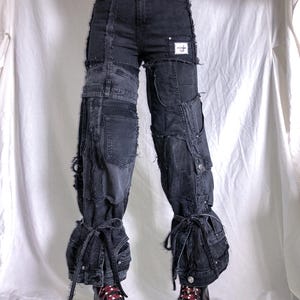 Könnte beinhalten: Dunkelgraue Jeanshose mit Patchwork-Design. Die Hose hat mehrere Taschen, ausgefranste Kanten und Bindebänder an den Knöcheln. Ein kleiner weißer Aufnäher ist am rechten Bein sichtbar. Die Hose wird mit schwarzen Plateauschuhen getragen.