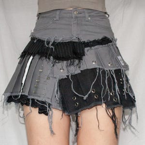 Op de afbeelding: Een grijze denim minirok met een gelaagd, geconstrueerd ontwerp. De rok heeft rafelige randen, metalen klinknagels en zwarte stoffen patches. De rok heeft een geplooide voorkant en een rauwe zoom.