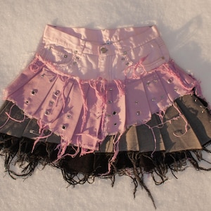Könnte beinhalten: Ein rosa Jeansrock mit einem mehrschichtigen Design. Der Rock hat ausgefranste Kanten, Metallösen und einen ausgefransten Saum. Der Rock hat ein plissiertes Design mit einer Mischung aus rosa, grau und schwarzen Stoffen.