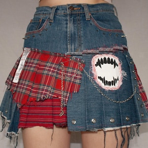Könnte beinhalten: Jeans-Minirock mit roten Tartan-Einsätzen und einem weißen und schwarzen Monsterzahn-Patch. Der Rock hat silberne Ösen, eine Kette und ausgefranste Kanten, die eine Punkrock-Ästhetik zeigen.