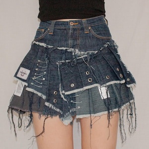 Könnte beinhalten: Dunkelblaue Jeansrock mit einem mehrschichtigen, verwitterten Design. Der Rock hat ausgefranste Kanten, Sicherheitsnadeln und Metallösen. Aufnäher aus hellerem Denim sind eingearbeitet.
