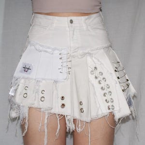 Può includere: Gonna di jeans bianca con un design decostruito. Presenta bordi strappati, spille da balia, occhielli e una toppa con una grafica. La gonna ha una costruzione unica e stratificata con un'estetica grezza e audace.