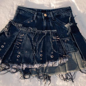 Può includere: Gonna in denim blu scuro con un design a strati e destrutturato. Presenta pieghe, occhielli metallici, spille di sicurezza e bordi sfilacciati. La gonna ha una chiusura con bottoni e tasche, mostrando uno stile unico e audace.