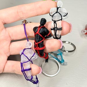 May include: A collection of keychains featuring small human-shaped figures in various colours, including clear, black, and blue, each bound in intricate patterns of coloured cord such as red, purple, black, and teal, attached to silver metal key rings.
