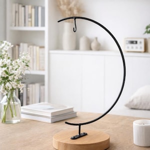 May include: A black metal display stand with a curved top and a hook, mounted on a round wooden base. The stand is on a light brown wooden table, with a white background.