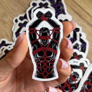 May include: A die-cut sticker featuring a stylized illustration of a figure with a blindfold and torso bound in intricate red rope patterns against a solid black background. The figure's arms are raised and crossed above the head, secured by rope.