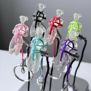 May include: A collection of six translucent frosted resin human-shaped keychains, each featuring a different colored cord tied in a decorative pattern around the torso. The keychains are attached to silver-toned metal rings and displayed on stands.