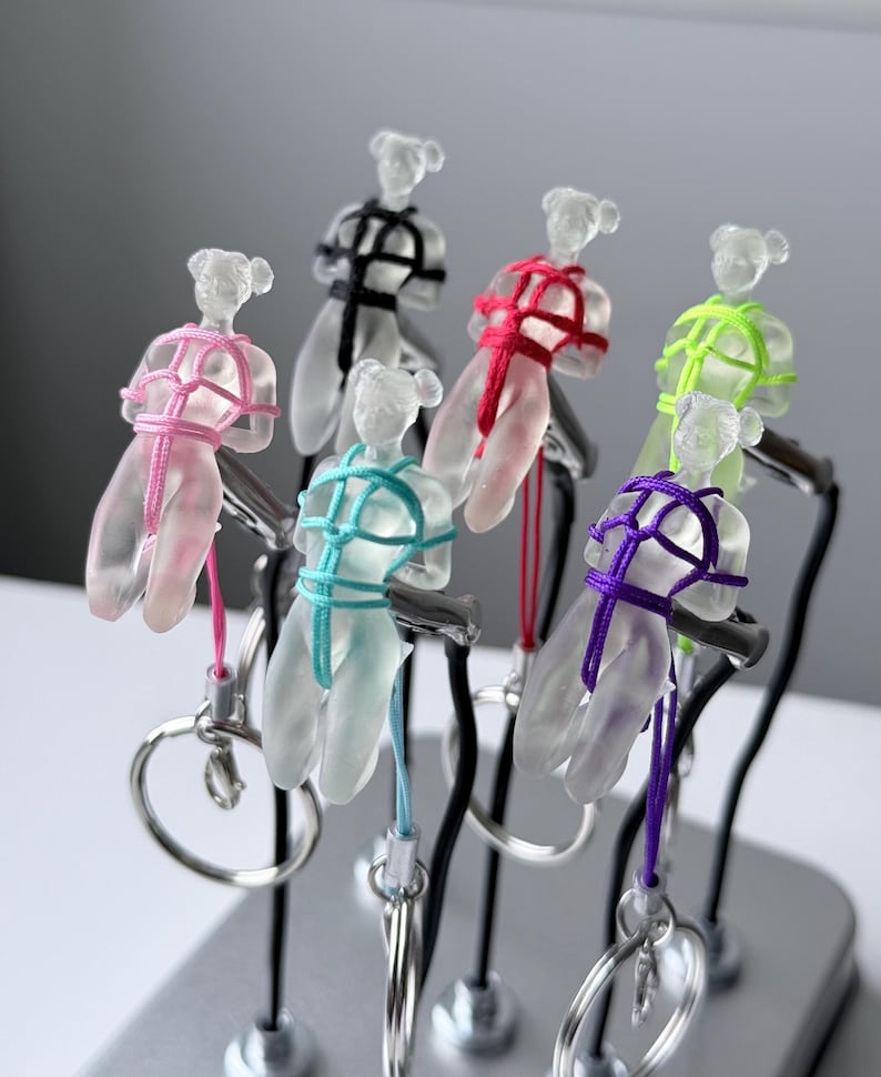 May include: A collection of six translucent frosted resin human-shaped keychains, each featuring a different colored cord tied in a decorative pattern around the torso. The keychains are attached to silver-toned metal rings and displayed on stands.
