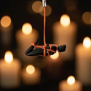 May include: A black figurine of a person in a prone position, bound with bright orange cord. The figurine is suspended from a silver-toned metal keyring by a vertical orange cord attached to the center of the binding, creating a hanging ornament.