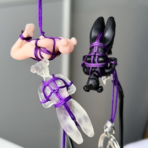 May include: Three small human-shaped figurines suspended by purple nylon cord. One figurine is matte black, one is translucent frosted glass, and one is a light peach color. Each figure is bound in a decorative knot pattern with the purple cord.