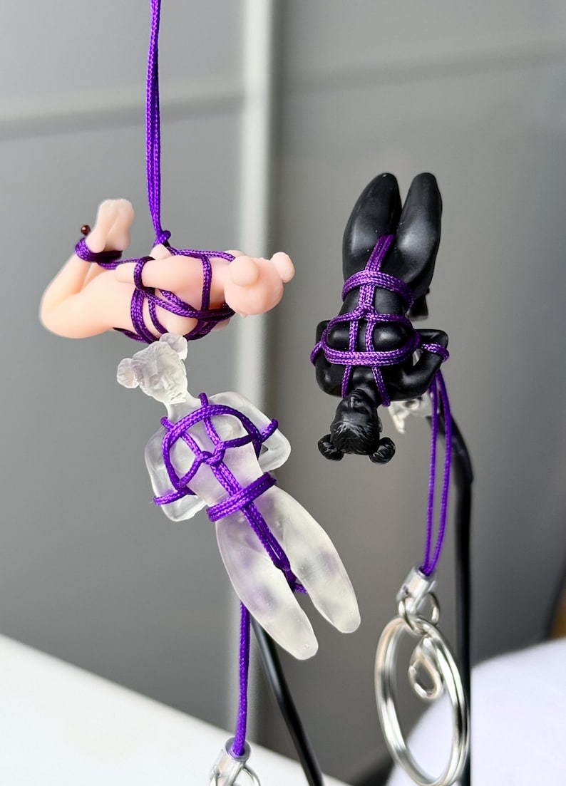 May include: Three small human-shaped figurines suspended by purple nylon cord. One figurine is matte black, one is translucent frosted glass, and one is a light peach color. Each figure is bound in a decorative knot pattern with the purple cord.