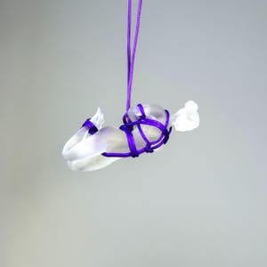 May include: A translucent, frosted glass figurine of a reclining figure suspended by a vibrant purple cord. The figure is bound with the same purple cord, which wraps around the torso and limbs, creating a decorative, knotted aesthetic.