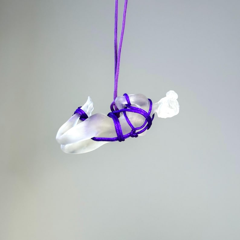 May include: A translucent, frosted glass figurine of a reclining figure suspended by a vibrant purple cord. The figure is bound with the same purple cord, which wraps around the torso and limbs, creating a decorative, knotted aesthetic.