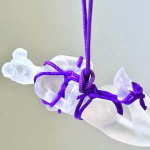 May include: A translucent, frosted glass figurine of a reclining figure suspended by a vibrant purple nylon cord. The cord is intricately wrapped around the torso and limbs of the glass sculpture, creating a series of knots that secure the piece.
