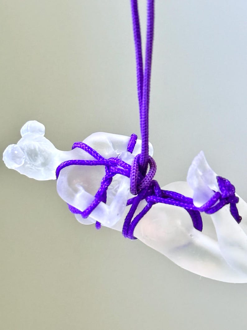 May include: A translucent, frosted glass figurine of a reclining figure suspended by a vibrant purple nylon cord. The cord is intricately wrapped around the torso and limbs of the glass sculpture, creating a series of knots that secure the piece.