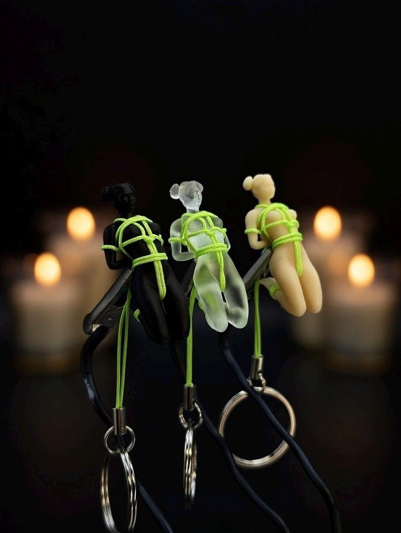 May include: Three small human-shaped figurines in black, translucent frosted, and beige colors, each bound with bright neon green cord. The figures are attached to black straps with silver-toned metal key rings hanging from the bottom of each piece.