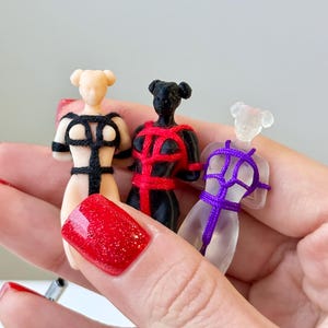 May include: Three small figurines of human torsos, each featuring a different color and material: a beige figure with black cordage, a black figure with red cordage, and a translucent clear figure with purple cordage, all tied in a harness pattern.