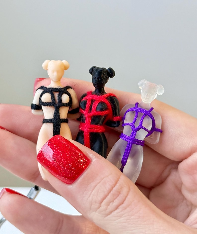 May include: Three small figurines of human torsos, each featuring a different color and material: a beige figure with black cordage, a black figure with red cordage, and a translucent clear figure with purple cordage, all tied in a harness pattern.
