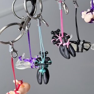 May include: A collection of keychains featuring small, stylized figures in black or flesh-toned colors, each bound with colorful cords in red, purple, light blue, pink, or black. The figures are suspended from silver-toned metal key rings.