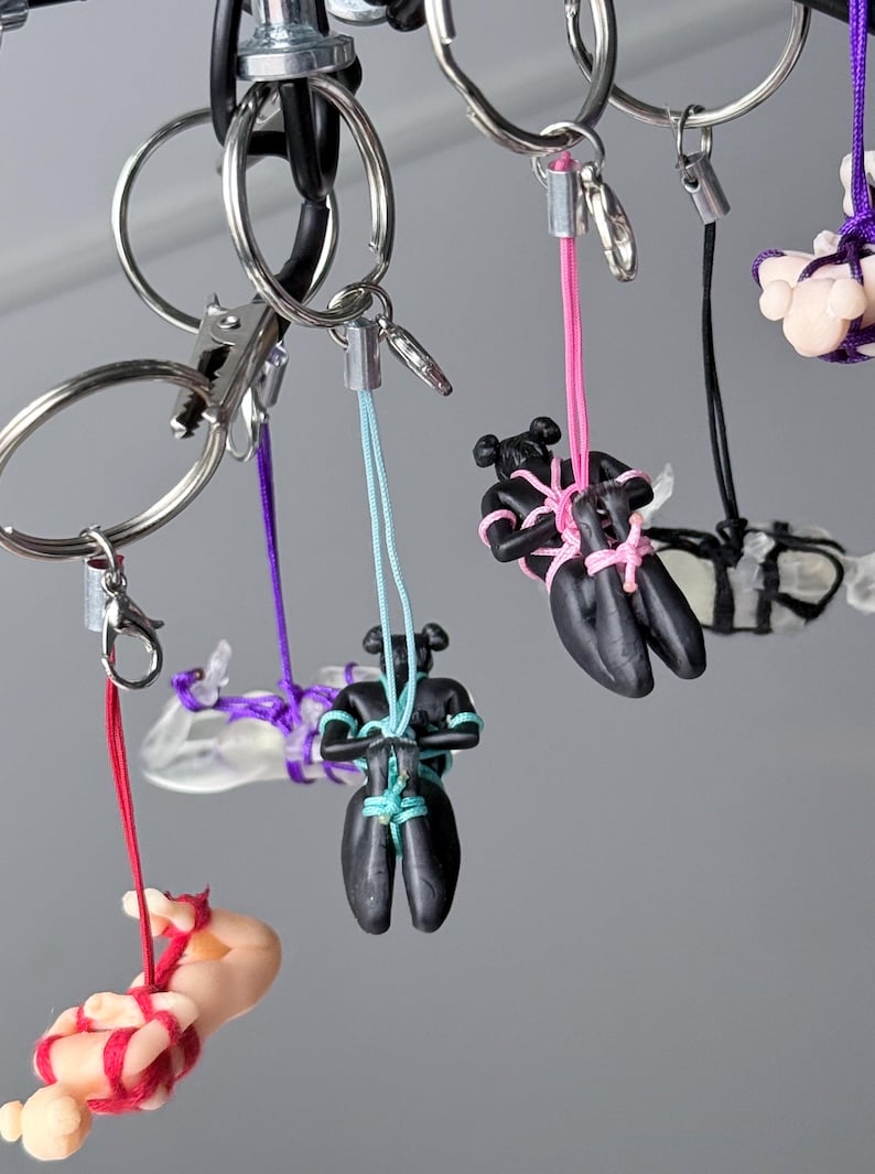 May include: A collection of keychains featuring small, stylized figures in black or flesh-toned colors, each bound with colorful cords in red, purple, light blue, pink, or black. The figures are suspended from silver-toned metal key rings.