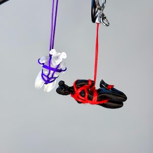 May include: Two hanging charms suspended from a black metal ring. One charm is a translucent white bear figure bound in purple cord. The second charm is a solid black human-like figure in a horizontal position, bound in bright red cord.