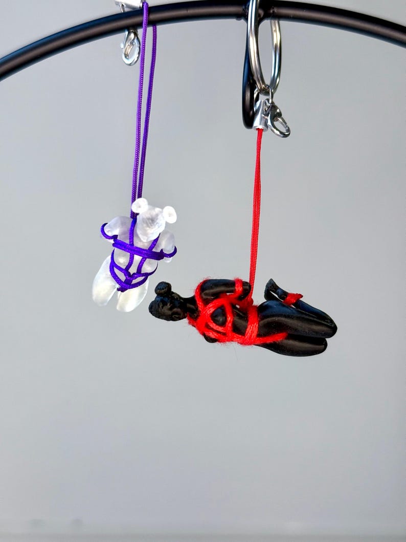 May include: Two hanging charms suspended from a black metal ring. One charm is a translucent white bear figure bound in purple cord. The second charm is a solid black human-like figure in a horizontal position, bound in bright red cord.