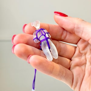 May include: A small, translucent, frosted glass figurine shaped like a human form, bound in a decorative pattern with a thin, vibrant purple cord. The cord extends downward from the figurine, ending in a small, metallic attachment component.