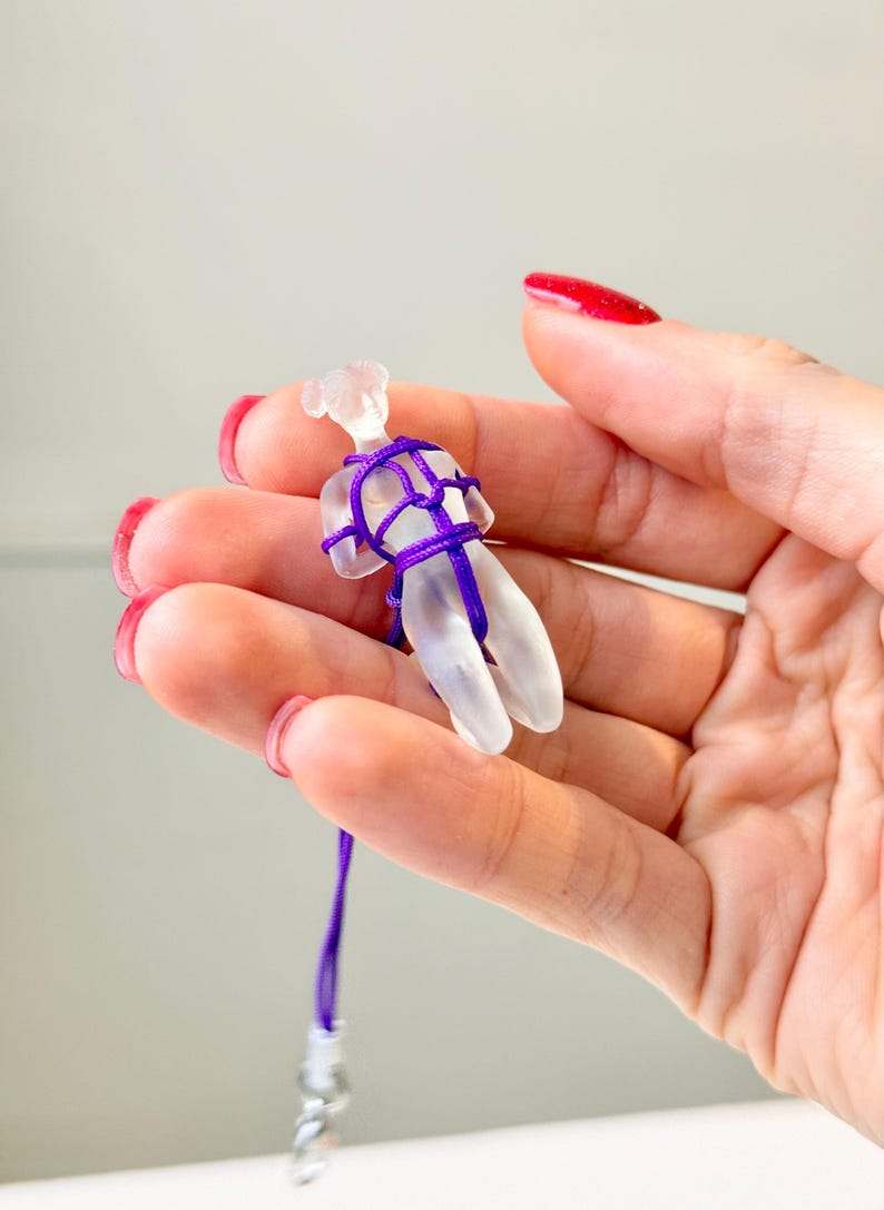 May include: A small, translucent, frosted glass figurine shaped like a human form, bound in a decorative pattern with a thin, vibrant purple cord. The cord extends downward from the figurine, ending in a small, metallic attachment component.