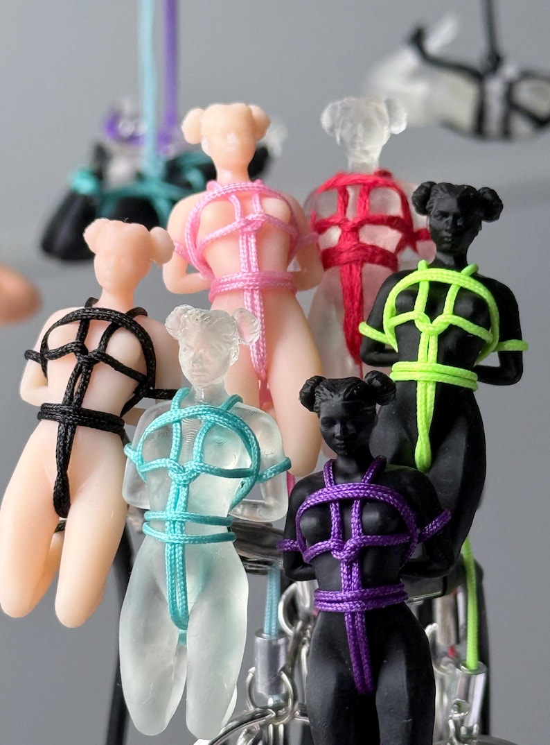 May include: A collection of small, human-shaped figurines in various colors including matte black, translucent clear, and peach. Each figure is bound in intricate, colorful nylon cord patterns, featuring neon green, bright pink, purple, black, and turquoise rope.