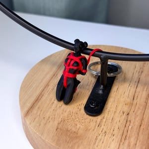May include: A black figurine of a person in a kneeling position, bound with bright red cord. The figurine is attached to a metal ring, which is suspended from a curved black metal bar mounted on a circular wooden base with a visible wood grain.