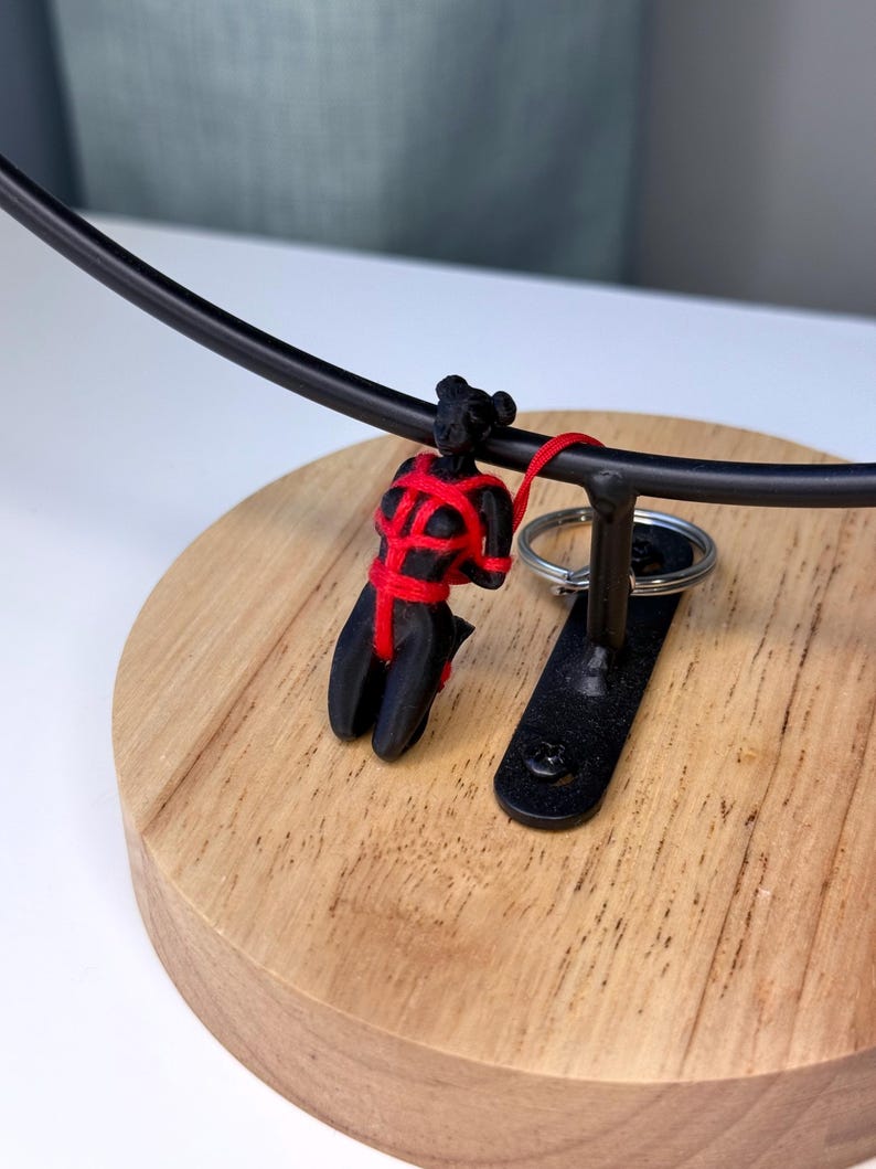 May include: A black figurine of a person in a kneeling position, bound with bright red cord. The figurine is attached to a metal ring, which is suspended from a curved black metal bar mounted on a circular wooden base with a visible wood grain.