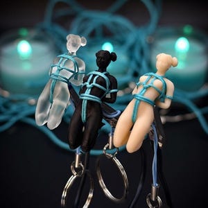 May include: Three small human-shaped figurines in clear, black, and beige colors, each bound in bright blue cord. The figures are attached to metal clips and silver-toned key rings. The figurines feature a smooth finish and a tied rope design.