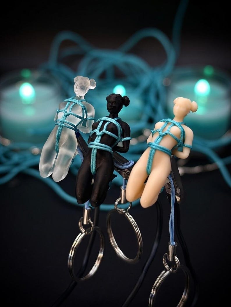 May include: Three small human-shaped figurines in clear, black, and beige colors, each bound in bright blue cord. The figures are attached to metal clips and silver-toned key rings. The figurines feature a smooth finish and a tied rope design.