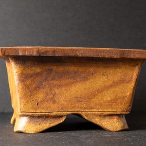 Könnte beinhalten: Ein rechteckiger, handgefertigter Bonsai-Topf in einem warmen, erdigen Braunton. Der Topf hat eine leicht strukturierte Oberfläche und wird von drei kleinen Füßen getragen. Der Rand ist dunkler braun.