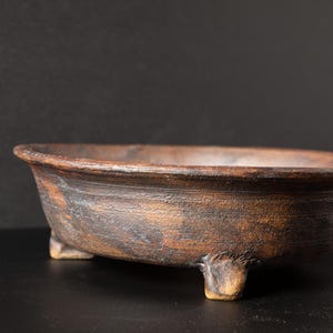 May include: A brown, oval-shaped ceramic bonsai pot with three small feet. The pot has a textured surface and a dark, earthy color, suggesting a natural, handcrafted aesthetic. The rim is slightly flared.
